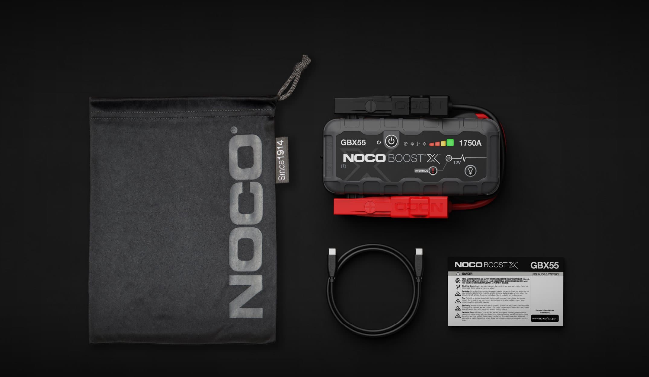 The NOCO Boost X GBX55 laid out with its contents: the jump starter with its attached red and black clamps, a USB-C to USB-C cable, a black NOCO microfibre storage bag and the safety and warranty booklet